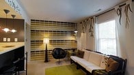 Located at 5400 Beverly Hills Dr in Columbus, Ohio, this single-floor residence offers a thoughtfully designed living space with a strong contemporary aesthetic. The living area immediately draws attention with a bold, geometric accent wall featuring a grid pattern in rich tones of gold, olive, and dark brown — a striking focal point that sets the tone for the entire space. Warm ambient lighting from sculptural floor lamps complements the natural light that flows in through large windows dressed with decorative curtains, creating a balanced and inviting atmosphere. The seating arrangement includes a sleek black leather swivel chair and a clean-lined white sofa with dark trim, reflecting a modern sensibility with contrasting textures and colors. A vibrant chartreuse area rug anchors the space and adds a lively pop of color against the neutral carpeted flooring. A small accent table rounds out the arrangement, offering both style and function. The overall ambiance is bold yet comfortable, combining graphic design elements with carefully chosen furnishings to create a space that feels curated and distinctive. Natural light and well-placed artificial lighting ensure the space feels bright and welcoming throughout the day.