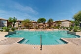 Located at 17374 N 89th Ave in Peoria, Arizona, this outdoor amenity area showcases a resort-style design rooted in Southwest Spanish architecture. The complex features multiple swimming pools of varying shapes and depths, catering to a range of recreational needs. Each pool is finished with decorative tile accents and surrounded by generously sized concrete sun decks lined with modern chaise lounges, providing ample space for relaxation under the Arizona sun.

A standout feature is the elevated spa, encircled by natural stone detailing and framed by desert-adapted plantings including palm trees, agave, and flowering shrubs. A central covered pavilion with a clay tile roof and arched entryway serves as a focal gathering point, offering shaded seating and views across the pool area.

The architecture throughout is consistent with a warm, stucco-clad aesthetic accented by terracotta roof tiles, creating a cohesive and visually inviting environment. Mature trees and manicured hedgerows provide both shade and privacy, enhancing the sense of a self-contained retreat. Iron perimeter fencing adds a layer of security while maintaining open sightlines to the surrounding landscaped grounds. The overall ambiance is that of a well-appointed, community-focused outdoor destination designed for year-round enjoyment in the desert climate.