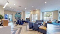 The lobby at 28 Southfield Ave in Stamford, Connecticut offers a polished and thoughtfully designed welcome experience. The space is anchored by a striking herringbone-patterned hardwood floor rendered in varying shades of grey, tan, and blue, lending the area a dynamic visual foundation. A coastal design theme runs throughout, most notably expressed through a large circular ocean mural that serves as a dramatic focal point on one wall, complemented by a decorative hanging wicker chair suspended beneath it.

The main seating area features an arrangement of modular blue upholstered sofas and ottomans, providing ample lounge space for residents and guests. A dedicated reception desk with clean-lined wood and light-toned finishes sits prominently in the lobby, accented by lush indoor greenery that adds warmth and life to the environment. Recessed lighting throughout the ceiling ensures even, bright illumination across the entire space.

A quieter sitting alcove opens off the main lobby, furnished with contemporary accent chairs arranged around a geometric wood coffee table, and framed by a large window that fills the nook with natural light and outdoor views. Contemporary wall art and sliding glass room dividers add refined detail, while the overall color palette of soft beige, white, and oceanic blue tones creates a calm, inviting atmosphere throughout.