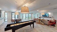 This single-floor amenity lounge is thoughtfully designed to serve as a multifunctional social hub, combining relaxed gathering areas with recreational features. The space is anchored by herringbone-patterned flooring that runs throughout, lending a refined yet casual coastal aesthetic. Shiplap-clad ceilings add architectural texture and warmth, while oversized woven pendant lights provide ambient illumination across the open layout.

A full-size shuffleboard table serves as a central recreational focal point, complemented by a dedicated game table surrounded by upholstered chairs — ideal for cards or board games. On one side of the room, a built-in lower cabinetry unit with a rich wood ledge shelf runs along the wall, accented by framed artwork and recessed lighting, creating a clean and organized backdrop.

The lounge area features a plush sectional sofa, cognac leather armchairs, and a rustic live-edge console table, all arranged around a wall-mounted flat-screen television. Decorative accents, including sculptural pieces and woven wall art, add personality throughout.

Floor-to-ceiling black-framed windows and glass doors line the perimeter, flooding the space with natural light and offering open views of an outdoor terrace and landscaped surroundings, seamlessly connecting the interior to the exterior environment.