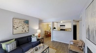 This single-floor, 2-bedroom, 2-bathroom residence in Harrisburg is designed for comfortable, everyday living with a thoughtful, open layout that seamlessly connects the main living areas. The living room serves as the heart of the home, featuring warm hardwood-style flooring, neutral gray walls, and a textured ceiling that adds subtle character. A large sectional sofa and coffee table anchor the space, which flows naturally into the adjacent dining area, defined by a pendant light fixture and wood-toned dining set. The kitchen is positioned just beyond, separated by a half-wall pass-through that maintains an open, airy feel throughout. It showcases white cabinetry, a classic subway tile backsplash, dark countertops, and black appliances, offering a clean, contemporary aesthetic. The pass-through design promotes connectivity between the kitchen and living spaces, making it ideal for entertaining or everyday interaction. Natural light filters through the space, enhancing the bright, welcoming atmosphere created by the light-toned walls and flooring. The overall ambiance is clean, modern, and functional — a well-appointed residence that balances practicality with a comfortable, inviting style suited to Harrisburg living.