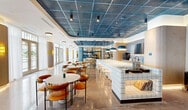 Located at 2250 NW 1st Ave in Miami's dynamic Wynwood neighborhood, this single-floor amenity space offers a thoughtfully designed, multi-functional environment spanning a generous footprint. The interior is defined by a striking blue metal mesh ceiling that anchors the main social area, complemented by polished terrazzo and concrete floors that flow throughout the space.

The café and bar area features a white tile-clad counter with bar seating, blue pendant lighting, and warm wood shelving, creating an inviting hub for residents and visitors. Adjacent to it, a sunlit lounge area is lined with floor-to-ceiling windows dressed in sheer white curtains, allowing abundant natural light and outdoor views. Unique suspended swing chairs, plush yellow sofas, and a billiards table add a playful, resort-like quality to the lounge.

A dedicated co-working and dining zone includes a large communal table with white Scandinavian-style chairs beneath modern pendant fixtures, set against a colorful area rug. Two private conference rooms offer focused meeting space — one featuring a bold geometric acoustic panel wall with an integrated display screen.

The game room rounds out the amenities with arcade machines, a large wall-mounted screen, and casual seating. Throughout, curated artwork, lush greenery, and a cohesive palette of blue, yellow, and warm wood tones create a vibrant, energetic atmosphere.