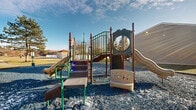 Located at 5179 Dempster Dr in Columbus, Ohio, this outdoor playground area offers a well-equipped recreational space designed for active play. The centerpiece of the area is a multi-component playground structure featuring multiple slides — including a curved slide and a straight slide — along with climbing stairs, railings, a circular mirror panel, and various interactive play elements. The structure is constructed with durable materials in earth tones of tan, green, and brown, giving it a sturdy and inviting appearance.

The play area is surfaced with blue rubber mulch/gravel, providing a safety-conscious ground cover that extends generously around the entire structure, offering ample open space for movement and play. The area is bordered by maintained green lawn, mature trees, and shrubs, contributing to a pleasant natural backdrop.

The surrounding environment includes multi-story residential buildings in close proximity, indicating this playground serves as a community amenity. The open layout allows for clear sightlines across the area, making supervision straightforward. Under clear skies, the space benefits from abundant natural light, creating a bright and welcoming outdoor environment suitable for children of various ages.