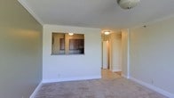 This single-floor residence offers a clean, classic interior characterized by crown molding, flush-mount ceiling lighting, and neutral-toned carpeting throughout the main living and sleeping areas. The living area connects visually to the kitchen via a pass-through window framed by a granite-topped counter, providing an open, communicative layout without fully removing the separation between spaces. The kitchen features dark wood cabinetry and appears to include standard appliances, including what appears to be a refrigerator visible through the pass-through opening. A hallway leads from the main living space toward additional rooms, with light tile or hardwood flooring transitioning from the carpeted area. The bedroom, accessible through a wide doorway, is a generously sized room with plush carpeting, crown molding, and a broad multi-pane window fitted with vertical blinds. The window allows considerable natural light to fill the space. A baseboard heating system runs along the perimeter of the bedroom. The overall aesthetic is clean and understated, with white walls and white trim creating a bright, neutral backdrop throughout.