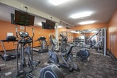 This fitness center is designed to support a full range of workout routines within a single, open-plan space. The interior is immediately distinguished by its warm orange walls, which lend an energetic atmosphere throughout. Patterned carpet tiles cover most of the floor, transitioning to rubber matting in the dedicated strength training zone, providing both comfort and safety during exercise.

The cardio area lines one side of the room and includes a row of elliptical machines and treadmills, each positioned beneath wall-mounted televisions for entertainment during workouts. Ceiling fans and recessed fluorescent lighting ensure the space remains well-lit and ventilated. A full-length mirror runs along one wall of the strength training section, visually expanding the room while allowing users to monitor their form.

The strength training area features multiple cable and resistance machines, adjustable benches, stability balls, and a dumbbell rack, offering versatility for various fitness goals. Colorful abstract artwork decorates the walls, adding a creative touch to the functional environment. Two entry points provide convenient access, with one door opening to an outdoor area and another connecting to an adjacent interior corridor. A wall clock and informational signage complete the practical amenities of this well-appointed fitness space.