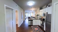 This single-story residence showcases an open-concept layout that seamlessly connects the kitchen and living areas, creating a cohesive and functional living environment. The kitchen is well-appointed with crisp white cabinetry, dark granite countertops, and a full suite of stainless steel appliances, including a dishwasher, range, and microwave. Pendant lighting hangs above the kitchen peninsula, providing both task lighting and a stylish focal point. Rich, dark hardwood-style flooring runs throughout the space, adding warmth and continuity. A decorative area rug anchors the kitchen zone, adding a pop of color. The open corridor leading toward the living area offers views of a bright, naturally lit sitting space, complete with a ceiling fan, patterned curtains, and a glass door that allows natural light to filter in. A hallway off the main corridor leads to a bathroom, suggesting a practical and efficient floor plan. The neutral wall tones complement the overall modern aesthetic, making the space feel clean, inviting, and move-in ready.
