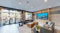 Located at 2501 N Orange Blossom Trail in Orlando, Florida, this generously sized single-floor community lounge offers a thoughtfully designed social space with a dynamic blend of modern and industrial aesthetics. The open-plan layout accommodates multiple distinct seating zones, each furnished with a curated mix of tufted leather sofas, vibrant accent chairs in orange, teal, and jewel tones, and contemporary coffee tables that create an inviting, eclectic atmosphere.

A standout focal point is the large, built-in aquarium set against a textured wall of reclaimed wood and exposed brick, adding a dramatic and lively visual element to the space. Rustic wooden columns rise from the polished tile flooring, reinforcing the industrial-chic design language throughout. Pendant lighting illuminates a bar-height communal dining and workspace area, accented by a decorative herringbone-patterned wall feature.

Floor-to-ceiling windows and sliding glass doors flood the interior with natural light while offering direct views and access to an outdoor patio area, complete with lounge seating surrounded by tropical Florida landscaping. A dual-sided fireplace with a sleek metal surround anchors the central seating area, adding warmth and architectural interest. Recessed lighting throughout ensures the space is well-lit at all times, completing this versatile and visually rich communal environment.