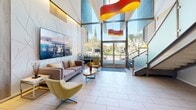 This 400 square foot space in Los Alamitos, CA, showcases a sleek, modern design with a focus on openness and light. The lobby area features high ceilings with a dramatic staircase, creating a sense of verticality and space. Large windows flood the area with natural light, offering views of the surrounding landscape. The color palette is predominantly neutral, with white walls and light-colored tile flooring, accented by pops of vibrant colors in the artwork and furnishings. Contemporary seating areas are strategically placed, featuring stylish sofas and chairs in muted tones. A striking sculptural element in red and yellow adds a dynamic visual focal point to the space. The decor is minimalist yet impactful, with carefully curated art pieces and potted plants adding warmth and texture. Recessed lighting and modern fixtures contribute to the overall ambiance. The space seamlessly blends functionality with aesthetic appeal, creating an inviting atmosphere for visitors and occupants alike.