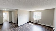 This single-floor unit is thoughtfully laid out to maximize functionality across its key living spaces. The open living area is finished with dark hardwood-style plank flooring that extends throughout, complemented by neutral gray walls and a textured popcorn ceiling. Natural light enters through a standard window offering outdoor views, while a wall-mounted HVAC unit and baseboard heating provide year-round climate control.

The kitchen features white cabinetry with sleek bar-pull hardware, a speckled gray laminate countertop, and a double stainless steel sink. A mosaic tile backsplash in muted gray and charcoal tones adds a contemporary accent to the space. Under-cabinet lighting enhances visibility and adds a practical touch.

The bedroom is carpeted in a soft, light gray pile and includes its own window that allows natural light to fill the room. A flush-mount ceiling light fixture provides overhead illumination, and baseboard heating runs along the exterior wall.

The bathroom is clean and functional, featuring a full-size soaking tub with an overhead shower and curtain rod, white subway tile surround, and a small window above the tub for ventilation and natural light. The overall aesthetic throughout the unit is neutral and move-in ready.