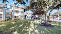 Located at 19100 Glenwest Dr in Houston, Texas, this residential community presents a welcoming presence defined by its distinctive teal and gray lap siding exteriors accented with crisp white trim and railings. The multi-story buildings are arranged to maximize access to lush, landscaped grounds shaded by large, mature oak trees that lend a well-established character to the property. Covered exterior corridors with tiled walkways lead to individual unit entrances, each marked with unit numbers and framed with white molding against the siding—offering both practicality and a cohesive aesthetic. The grounds are thoughtfully appointed with resident-focused amenities, including a children's playground area visible from the green lawn spaces that extend between buildings. A dedicated pet station with waste disposal is conveniently located near a decorative wrought-iron and stone-pillar fence line that borders the perimeter, offering open views beyond the property. Stairwells with metal railings connect the building levels, featuring the same signature teal siding throughout. Fire safety equipment is visibly installed along the exterior corridors. The overall ambiance is that of a community-oriented setting, combining functional design with green outdoor spaces ideal for everyday living in the suburban Houston area.