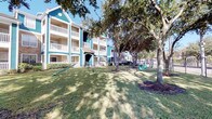 Located at 19100 Glenwest Dr in Houston, Texas, this residential community presents a welcoming presence defined by its distinctive teal and gray lap siding exteriors accented with crisp white trim and railings. The multi-story buildings are arranged to maximize access to lush, landscaped grounds shaded by large, mature oak trees that lend a well-established character to the property. Covered exterior corridors with tiled walkways lead to individual unit entrances, each marked with unit numbers and framed with white molding against the siding—offering both practicality and a cohesive aesthetic. The grounds are thoughtfully appointed with resident-focused amenities, including a children's playground area visible from the green lawn spaces that extend between buildings. A dedicated pet station with waste disposal is conveniently located near a decorative wrought-iron and stone-pillar fence line that borders the perimeter, offering open views beyond the property. Stairwells with metal railings connect the building levels, featuring the same signature teal siding throughout. Fire safety equipment is visibly installed along the exterior corridors. The overall ambiance is that of a community-oriented setting, combining functional design with green outdoor spaces ideal for everyday living in the suburban Houston area.