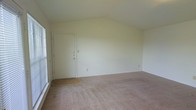This single-story space is characterized by its vaulted ceiling, which adds a sense of vertical dimension and openness to the interior. The main living area features wall-to-wall neutral-toned carpeting that extends throughout, while a tiled or hardwood entry area near the front door provides a practical transition zone upon entry. The front door is solid white and fitted with multiple locks, contributing to the security of the space. Walls are finished in a clean, off-white tone that reflects light and keeps the atmosphere feeling bright and airy. A window positioned along one side of the room allows natural light to filter in, further enhancing the open feel of the space. The vaulted ceiling rises toward the center of the room, creating an architectural focal point that distinguishes the space from standard flat-ceiling layouts. The overall design is simple and functional, presenting a blank slate well-suited for personalization. The layout is open and unobstructed, allowing for flexible furniture arrangement throughout the main living area.