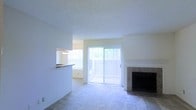 This single-floor residence offers a clean, open layout filled with natural light. The living area serves as the heart of the home, anchored by a tile-surround fireplace that adds a classic, functional focal point to the room. Soft carpeting covers the living space floor, contributing to a comfortable and welcoming atmosphere. Large sliding glass doors dressed with vertical blinds open to an outdoor balcony, allowing abundant natural light to flow into the interior and providing a seamless connection to the outdoors. Adjacent to the living area, a pass-through counter connects to the kitchen, enhancing the open flow between spaces and making the layout feel more expansive. The kitchen area, visible through the pass-through, benefits from its own window, further brightening the interior. Walls are finished in neutral tones throughout, complemented by a textured ceiling that adds subtle character. The overall design is straightforward and functional, with an emphasis on light, openness, and ease of movement between the living and kitchen areas. The space presents a practical yet comfortable single-level layout well-suited for everyday living.