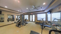 This fitness center offers a dedicated, single-floor space designed to support a well-rounded exercise routine. The room is thoughtfully laid out to accommodate both strength training and cardiovascular equipment, including a multi-station cable machine, weight benches, and treadmills positioned to allow comfortable use without crowding. Large windows fitted with blinds line the walls, allowing natural light to fill the space while maintaining privacy. A set of glass-paneled double doors provides convenient access and adds to the open, airy feel of the room. The ceiling features elegant tray detailing trimmed in dark brown molding, which adds a refined architectural touch to the otherwise functional space. Multiple ceiling fans are mounted throughout, ensuring consistent air circulation during workouts. Recessed lighting supplements the natural light, keeping the room well-lit at all times. Full-length mirrors along one wall visually expand the space and allow users to monitor their form during exercise. The light-colored carpeted flooring and neutral warm-toned walls contribute to a clean, welcoming atmosphere. Overall, this fitness center balances practicality with comfort, providing an inviting environment for daily exercise.