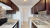 This single-floor space showcases a classic galley-style kitchen designed for functional efficiency. The layout features cabinetry running along both walls, finished in a rich, dark espresso tone with sleek silver bar-pull hardware. The upper and lower cabinets provide generous storage capacity, complemented by an integrated microwave above the range on one side.

The countertops are finished in a light granite or granite-style surface, offering a bright contrast against the darker cabinetry. A decorative mosaic tile backsplash runs beneath the upper cabinets on both sides, adding visual texture with its mixed stone-and-glass pattern in neutral tones.

Appliances include a white gas range and a white refrigerator on the left wall, while the right wall features a stainless steel sink with a single-handle faucet and a dishwasher integrated beneath the counter. The light ceramic tile flooring ties the space together with a clean, neutral finish.

Overhead, a flush-mount ceiling light provides even illumination throughout the corridor. At the far end, a doorway opens to an adjacent room, enhancing the sense of flow and connectivity within the space.