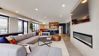 This thoughtfully designed single-floor amenity space blends contemporary style with functional versatility across several distinct zones. The main lounge area serves as the centerpiece, anchored by a generously sized sectional sofa with colorful accent pillows, a round leather ottoman, and a low-profile coffee table set atop a woven area rug. A sleek, tile-surround fireplace adds warmth along one wall, while floor-to-ceiling windows on multiple sides flood the space with natural light and provide sweeping outdoor views, including access to an exterior terrace with additional seating.

The kitchen area features dark navy cabinetry, a stacked brick accent wall, stainless steel appliances, a large island with bar seating, and pendant lighting overhead. A cozy built-in booth with orange leather upholstery provides casual dining seating nearby.

A separate meeting or dining room offers a striking design contrast, with bold teal walls, a reclaimed wood accent wall, a solid wood communal table surrounded by leather and metal chairs, and a statement multi-bulb chandelier. Curated artwork and built-in shelving complete the space, creating a refined yet welcoming environment suited for both collaboration and relaxation.