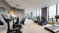Located at 500 Upper Wellington St in Hamilton, Ontario, this fitness facility offers a generously sized, single-floor exercise room designed to support a comprehensive workout experience. The space is defined by its floor-to-ceiling windows along one wall, which flood the interior with natural light and provide open views of the surrounding cityscape, creating an energizing atmosphere for exercise.

The room features a neutral color palette with light grey walls, recessed ceiling lighting, and durable, cushioned flooring throughout—practical for both cardio and strength training activities. A row of TRUE-brand treadmills and elliptical machines lines the windowed wall, maximizing the outdoor views during cardio sessions. Stationary bikes are positioned along the opposite side, complemented by a large mirrored wall that visually expands the space.

The strength training area includes a multi-function cable machine, adjustable weight benches, a barbell rack, and a tiered dumbbell rack offering a full range of free weights. A water fountain station is conveniently integrated into the layout. The overall design is clean, modern, and functional, providing a well-rounded facility suited for a variety of fitness routines.