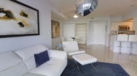This single-floor space showcases a cohesive, modern interior design anchored by a crisp white and navy color palette. The living area is furnished with sleek white leather seating, including a sofa accented with a deep navy cushion and a classic Barcelona-style chair paired with a matching ottoman, lending the space a sophisticated, mid-century modern aesthetic. A small circular glass side table adds a delicate touch to the seating arrangement.

A standout feature is the distinctive mirrored spherical pendant light, which serves as an eye-catching focal point and adds a bold, contemporary flair to the room. A traditional pendant light nearby provides complementary illumination. The flooring transitions between a deep navy area rug in the living zone and light-toned hard flooring throughout the remainder of the space.

Adjacent to the living area, a glass-top dining table with white chairs sits comfortably near built-in shelving and cabinetry finished in white, offering both display and storage functionality. Framed abstract artwork on the walls adds visual depth and character. The overall ambiance is clean, stylish, and well-composed, reflecting a polished contemporary aesthetic.
