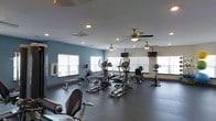 This single-floor fitness center offers a dedicated and well-appointed workout environment designed to support a range of cardio exercise routines. The space is outfitted with multiple elliptical trainers, treadmills, and stationary bikes, all arranged to allow comfortable movement between equipment. Dark rubber flooring covers the workout area, providing both durability and traction underfoot. The room is finished with a combination of cool blue and neutral cream-toned walls, lending the space a clean and energizing atmosphere. Natural light streams in through several windows fitted with white horizontal blinds, enhancing the open and airy feel of the interior. Multiple ceiling fans are installed throughout, promoting air circulation during workouts and contributing to the overall comfort of the space. Recessed lighting supplements the natural light, ensuring consistent illumination at all times of day. Wall-mounted flat-screen televisions are positioned at strategic points, allowing exercisers to stay entertained during their sessions. A defibrillator unit is also mounted on the wall, reflecting a commitment to safety. Overall, the fitness center is thoughtfully arranged to provide a functional, comfortable, and well-lit environment for cardiovascular training.