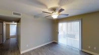 This single-story space offers a straightforward and functional layout designed for comfortable everyday living. The main living area is finished with hardwood-style flooring in a warm, neutral tone that complements the light gray walls and crisp white baseboards throughout. A ceiling fan with a built-in light fixture is centrally mounted on the textured ceiling, providing both air circulation and ambient lighting. A large sliding glass door fitted with vertical blinds serves as the room's primary source of natural light, brightening the interior and offering direct access to the outdoors. The textured ceiling adds a classic residential character to the space. The overall design is clean and uncluttered, with neutral tones throughout that offer a versatile backdrop for a variety of furnishing styles. The combination of natural light from the sliding door, durable flooring, and the ceiling fan contributes to a practical and comfortable atmosphere suited for everyday use.