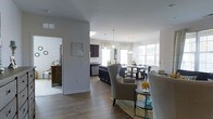 This single-floor residence showcases an open-concept layout that elegantly connects the living, dining, and kitchen areas into one cohesive, flowing space. The interior is finished with warm hardwood-style flooring that runs throughout, complementing the crisp white walls and creating a clean, contemporary aesthetic.

The kitchen features dark cabinetry that provides a striking contrast against the light surroundings, and is equipped with modern appliances. Pendant lighting hangs above the kitchen area, adding both task illumination and a stylish focal point. The space transitions naturally into a dedicated dining area, which is bathed in natural light streaming through a series of large windows. A chandelier anchors the dining space, adding an elegant touch.

The living area is generously proportioned, furnished with comfortable seating including wingback chairs and a sofa, centered around a area rug that defines the space. An adjacent room, visible through an open doorway, offers additional flexible living or bedroom space with its own natural light source.

Throughout the home, the open sightlines and abundant window placement create a bright, airy ambiance. The thoughtful combination of dark and light elements, along with consistent flooring, gives the entire space a cohesive and well-designed character.