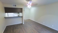 This single-floor space offers a straightforward, functional layout centered around an open living area with rich, dark hardwood-style flooring that extends throughout. The main living space is well-proportioned, featuring neutral beige walls, clean white baseboards, and a ceiling fan with an integrated light fixture that provides both ambient lighting and air circulation. A textured ceiling adds subtle visual interest overhead, while a wall-mounted thermostat indicates climate control convenience.

The kitchen is partially separated from the living area by a half-wall partition, creating a pass-through design that maintains an open feel while defining the cooking space. The kitchen features white appliances, including a range, along with a countertop that extends to form a breakfast bar along the partition. Dark cabinetry above the pass-through provides overhead storage and creates a visual contrast against the lighter walls. A sink with a standard faucet is visible along the kitchen's back wall. The overall design is clean and practical, with a neutral color palette that provides a versatile backdrop throughout the space.