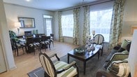 This well-appointed single-floor residence offers an inviting open-concept layout that seamlessly connects its living and dining areas. The space is anchored by warm hardwood flooring that runs throughout, lending a sense of continuity and warmth to the interior. Large windows dressed with elegant floral-patterned curtains flood the space with abundant natural light, creating an airy and welcoming atmosphere. The dining area features a dark wood table paired with upholstered chairs, offering a refined setting for meals and gatherings. A decorative mirror adorns the wall nearby, enhancing the sense of depth and brightness in the room. The sitting area showcases a dark wood accent table styled with silver candelabras and a decorative bowl, flanked by striped armchairs that add a classic, tailored touch. The neutral color palette of soft grays and greens, complemented by rich dark wood furnishings, creates a balanced and sophisticated aesthetic. An entry door with a sidelight window provides a welcoming entry point, while the generous window placement throughout ensures the space remains bright and connected to the outdoors. Overall, the space combines functionality with understated elegance, making it both practical and visually appealing.