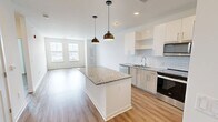 Located at Kingston Collection in Kingston, MA, this well-appointed one-bedroom, one-bathroom residence offers a thoughtfully designed single-floor layout with clean, modern finishes throughout. The open-concept kitchen and living area serves as the heart of the home, showcasing light wood cabinetry, a granite-topped central island with pendant lighting, subway tile backsplash, stainless steel appliances, and recessed lighting — all set atop warm hardwood-style flooring that flows seamlessly through the space. Large windows dressed with horizontal blinds invite generous natural light into the living area, creating a bright and airy atmosphere. The bedroom is comfortably sized, featuring dual windows with outdoor views, a ceiling fan, and the same warm flooring found throughout. A dedicated walk-in closet with wire shelving provides ample storage. The bathroom is well-appointed with a granite-topped vanity, wood-toned cabinetry, a framed mirror with accent lighting, and a tiled shower-tub combination. Clean white walls and crisp trim work are consistent throughout, lending the entire residence a fresh, contemporary character well-suited for modern living.