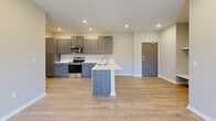 This single-floor unit offers a clean, modern design with a neutral color palette and thoughtful layout throughout. The open-concept living and dining area is anchored by smooth light-toned luxury vinyl plank flooring, recessed lighting, and a textured ceiling. A large front-facing window and glass-panel entry door allow natural light to fill the space, while an open passage connects seamlessly to additional rooms.

The kitchen is a standout feature, equipped with stainless steel appliances including a range, microwave, and refrigerator. Two kitchen configurations are visible—one with rich dark-toned cabinetry paired with a peninsula countertop and single-basin sink, and another with gray-toned cabinetry surrounding a central island with a sink. Both offer ample storage and bright white countertops that contrast elegantly with the cabinetry.

The bedrooms are finished with soft neutral carpet and feature windows that bring in outdoor views. A digital thermostat is conveniently mounted in one of the living areas. The bathroom includes tile flooring, a dark wood vanity with a mirror and vanity lighting, and a walk-in shower. A dedicated laundry room houses a full-size washer and dryer with overhead wire shelving for added storage.