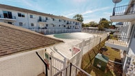 This single-floor residential unit is situated within a multi-unit building complex that offers a range of functional outdoor amenities. The exterior of the unit is clad in classic white vinyl horizontal siding, which gives the building a clean, uniform appearance. A covered patio area extends from the unit, featuring a lattice overhead structure that provides partial shade while maintaining an open, airy feel. The patio is accessible through a sliding screen door, offering a practical transition between the interior living space and the outdoors. Windows along the exterior wall are fitted with horizontal blinds, allowing for adjustable privacy and light control.

One of the standout features of this property is access to a shared outdoor pool area, enclosed by a white vinyl privacy fence with a gated entry. The pool is fitted with a safety cover and is set within a spacious concrete deck that invites outdoor relaxation. Informational signage is posted at the entrance, and the surrounding area is well-maintained. The broader complex is a two-story building with balconies visible along the upper level, set against a backdrop of mature trees and an open sky. The combination of private patio access and shared pool facilities makes this a well-rounded residential offering suited for comfortable everyday living.