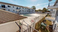 This single-floor residential unit is situated within a multi-unit building complex that offers a range of functional outdoor amenities. The exterior of the unit is clad in classic white vinyl horizontal siding, which gives the building a clean, uniform appearance. A covered patio area extends from the unit, featuring a lattice overhead structure that provides partial shade while maintaining an open, airy feel. The patio is accessible through a sliding screen door, offering a practical transition between the interior living space and the outdoors. Windows along the exterior wall are fitted with horizontal blinds, allowing for adjustable privacy and light control.

One of the standout features of this property is access to a shared outdoor pool area, enclosed by a white vinyl privacy fence with a gated entry. The pool is fitted with a safety cover and is set within a spacious concrete deck that invites outdoor relaxation. Informational signage is posted at the entrance, and the surrounding area is well-maintained. The broader complex is a two-story building with balconies visible along the upper level, set against a backdrop of mature trees and an open sky. The combination of private patio access and shared pool facilities makes this a well-rounded residential offering suited for comfortable everyday living.
