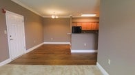 This single-floor space showcases a thoughtfully designed open layout that seamlessly connects the living and kitchen areas. The living and dining space features rich, dark hardwood flooring that contrasts elegantly with the warm taupe-toned walls and crisp white baseboards. Crown molding runs along the ceiling throughout, adding a refined architectural detail that elevates the overall aesthetic. A classic chandelier serves as a focal point in the dining area, providing warm ambient lighting.

The kitchen is positioned at the rear of the main living space and is defined by a raised breakfast bar counter, offering both a functional workspace and a natural division between areas. Warm honey-toned wood cabinetry lines the upper kitchen walls, complemented by dark countertops and black appliances. A decorative column anchors the transition between the kitchen and living space, adding a structural design element. Recessed and flush-mount ceiling lighting in the kitchen ensures the area is well-lit. Plush carpeting is visible at the foreground, suggesting an adjacent room or entryway zone, adding to the layered flooring design of the space.