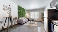This efficiently designed single-floor unit at Onyx makes a bold first impression with its striking floor-to-ceiling living green wall, which serves as the focal point of the combined kitchen and living area. The lush botanical feature wall, branded with the Onyx name, brings a vibrant, organic energy to the space and pairs beautifully with the neutral gray tones and soft carpeting throughout the living zone. A large window floods the living area with natural light and offers views of an outdoor seating area, enhancing the open and airy feel of the room. The kitchen is thoughtfully integrated into the same space, featuring dark espresso cabinetry, stainless steel appliances including a refrigerator, range, and microwave, and a clean white countertop with a modern sink. Wood-look flooring in the kitchen area transitions seamlessly into the carpeted living space. A short hallway connects the main living area to the bathroom, and features textured walls and decorative artwork. The bathroom is finished with wood-look tile flooring, a sleek vessel sink atop a dark vanity, a framed mirror, and a shower-tub combination, offering a clean and functional design. The overall ambiance is modern, cozy, and thoughtfully curated.