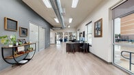This single-floor commercial space combines modern industrial design with a warm, functional layout. The entry reception area features large floor-to-ceiling windows that flood the space with natural light, a front desk with seating, and views of the exterior. Light wood-look flooring runs consistently throughout, complementing the neutral cream and bold slate-gray accent walls.

A prominent design element is the exposed metallic ductwork that runs along the ceiling, contributing to an industrial-modern aesthetic. The main corridor leads past frosted-glass sliding doors that open to private office or service rooms, with motivational wall decals adorning the gray feature wall. A bulletin board area and multifunction printer station are conveniently positioned along the hallway.

The open lounge area features a comfortable blue sectional sofa, wall-mounted television, and lush greenery displays — creating a relaxed, inviting atmosphere. Adjacent to the lounge is a fully equipped kitchen with granite countertops, a bar-height island with stools, light upper cabinetry, dark lower cabinetry, pendant lighting, and black appliances including a refrigerator and range. The space also includes private rooms with glass-panel doors, providing flexible use for meetings or individual workspaces throughout.