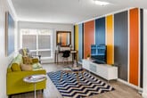 Located in the heart of Santa Ana, CA, this single-floor residence offers a thoughtfully designed layout with a cohesive mid-century modern aesthetic. The open-concept kitchen is equipped with warm wood cabinetry, granite-style countertops, black appliances including a refrigerator and range, and a peninsula with bar seating that connects seamlessly to the living and dining areas. Floating shelves provide decorative display space just off the kitchen entry. The living area is bright and welcoming, featuring wood-look flooring, a large window that allows generous natural light, and a bold accent wall with colorful vertical stripes that adds a distinctive visual element to the space. The bedroom is generously sized with plush carpeting, a striking deep-blue feature wall, and a sliding glass door that opens directly to a private outdoor patio. The bathroom includes a wood-vanity with granite-style countertop, a large mirror with overhead lighting, and a combined tub and shower with glass sliding doors. Throughout the residence, the design maintains a consistent palette of warm wood tones, bold accent colors, and clean white walls, creating an energetic yet inviting atmosphere.