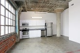 This single-floor space showcases a distinctive industrial aesthetic rooted in its original architectural character. Polished concrete floors run throughout, providing a durable and visually cohesive foundation. Exposed red brick walls serve as a defining feature, adding warmth and texture that contrasts with the smooth, neutral-toned drywall surfaces found in other areas.

The open main area benefits from generous ceiling height, with exposed ductwork, structural beams, and conduit left visible overhead, reinforcing the raw, loft-like character of the space. A structural column with a painted red base anchors the larger open zone, which offers substantial room for flexible use.

The kitchen is equipped with white cabinetry, stainless steel appliances including a refrigerator, dishwasher, electric range, and microwave, along with a double sink and dark countertops. Overhead fluorescent lighting illuminates the kitchen area effectively.

The bathroom features a walk-in shower with a white surround, a toilet, and a wood-fronted vanity with a countertop sink. Concrete floors continue into this room, maintaining material consistency throughout.

Additional interior spaces include rooms with closet storage and natural wood doors, contributing a warmer finish detail amid the otherwise utilitarian palette.