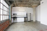This single-floor space showcases a distinctive industrial aesthetic rooted in its original architectural character. Polished concrete floors run throughout, providing a durable and visually cohesive foundation. Exposed red brick walls serve as a defining feature, adding warmth and texture that contrasts with the smooth, neutral-toned drywall surfaces found in other areas.

The open main area benefits from generous ceiling height, with exposed ductwork, structural beams, and conduit left visible overhead, reinforcing the raw, loft-like character of the space. A structural column with a painted red base anchors the larger open zone, which offers substantial room for flexible use.

The kitchen is equipped with white cabinetry, stainless steel appliances including a refrigerator, dishwasher, electric range, and microwave, along with a double sink and dark countertops. Overhead fluorescent lighting illuminates the kitchen area effectively.

The bathroom features a walk-in shower with a white surround, a toilet, and a wood-fronted vanity with a countertop sink. Concrete floors continue into this room, maintaining material consistency throughout.

Additional interior spaces include rooms with closet storage and natural wood doors, contributing a warmer finish detail amid the otherwise utilitarian palette.