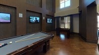 This single-floor game room is a well-appointed entertainment space defined by its dramatic soaring ceilings and upscale design details. The centerpiece of the room is a full-size billiard table finished in rich dark wood, complemented by a foosball table nearby, making the space well-suited for social gatherings and recreational use.

The walls feature a striking combination of dark wood paneling and textured stone-like surfaces, lending a refined, contemporary feel throughout. Two wall-mounted flat-screen televisions are positioned for easy viewing from multiple points in the room. Along one wall, a built-in counter with a row of modern swivel bar stools is backed by a bold, lattice-style decorative partition that serves as an eye-catching architectural focal point, filtering natural light from adjacent windows.

Large windows dressed with horizontal blinds allow natural light to fill the upper portions of the space, highlighting the vertical scale of the room. Warm hardwood flooring runs throughout, adding to the inviting atmosphere. Oversized framed mirrors are mounted on the walls, further enhancing the sense of depth and openness. A slim console table with decorative accessories adds a polished finishing touch to the overall design.