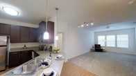This single-floor space showcases an open-concept layout that connects the kitchen and living area in a cohesive, functional design. The kitchen features dark espresso cabinetry that contrasts elegantly with light granite countertops, offering generous prep and storage space. A stainless steel sink and appliances add a modern, polished touch. Above the peninsula-style counter, two pendant lights with frosted glass shades provide warm, focused illumination, making the space ideal for casual dining or entertaining.

The transition from kitchen to living area is smooth, with hardwood-style flooring in the kitchen giving way to plush carpeting in the living room. A track lighting fixture mounted on the ceiling provides adjustable, ambient lighting throughout. The living area is bright and airy, benefiting from natural light filtering through a window dressed with blinds. White walls throughout the space enhance the sense of openness and provide a neutral backdrop that complements the darker kitchen elements. The overall ambiance is modern and welcoming, with a practical yet stylish design suited for everyday living.