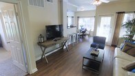 This single-floor residence offers an open-concept layout that connects the living, dining, and entertaining areas in one cohesive and well-lit space. Rich, dark hardwood-style flooring runs throughout, grounding the interior with a warm and inviting aesthetic.

The living area is thoughtfully arranged, featuring a media console with a large flat-screen television and a comfortable seating area anchored by a sleek, modern coffee table. Crown molding along the ceiling adds a refined architectural touch, while a ceiling fan contributes both style and functionality.

Flowing naturally from the living space, a bar-height counter with metal stools serves as a casual dining or workspace option, creating a smooth transition into the dedicated dining area beyond. The dining space accommodates a full table and chairs, bathed in natural light streaming through multiple windows dressed with dark curtain panels.

The abundance of windows throughout the space ensures generous natural light, enhancing the open and airy atmosphere. Neutral wall tones complement the dark flooring, creating a balanced contrast that feels both modern and timeless. The overall layout is practical and versatile, making excellent use of the available space while maintaining a clean, contemporary character throughout.