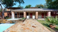 This single-story unit is part of a well-established brick building community set amid mature trees, with a charming stone-paved walkway leading to the entrance. The exterior features a covered porch with decorative columns, red accent doors, and a neighboring water feature that adds to the welcoming atmosphere of the grounds.

Inside, the layout is straightforward and functional. The living area is generously sized, finished with wall-to-wall carpet and a textured ceiling, offering a comfortable and quiet feel. Natural light filters in through a window fitted with blinds, brightening the open interior. The space flows easily between the living and bedroom areas, both of which share the same neutral-toned carpeting.

The kitchen is equipped with warm wood cabinetry, light-colored countertops, a double stainless steel sink, a white electric range, and a dishwasher. The hardwood-style flooring in the kitchen provides a practical contrast to the carpeted rooms.

The bathroom is clean and well-appointed, featuring a tub-shower combination with white tile surround, hardwood-style flooring, a vanity with wood cabinetry, light countertops, and a large mirror. Grab bars add an element of accessibility and practicality to the space.