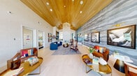 Located at 6741 Boulder Hwy in Las Vegas, Nevada, this single-floor community clubhouse at PARQ offers a generously sized, thoughtfully designed gathering space that blends vibrant contemporary style with practical amenities. The centerpiece of the space is a dramatic vaulted wood-paneled ceiling that rises to impressive heights, anchored by a striking sculptural gold chandelier that adds warmth and elegance throughout. Light-toned wide-plank flooring runs throughout, complementing the airy, open layout.

The main lounge area features a curved navy sectional sofa, richly upholstered leather accent chairs in cognac tones, and layered area rugs, all arranged around a modern fireplace set into a textured tile feature wall. A large-scale abstract wallcovering in cool grey tones serves as a dramatic backdrop for curated framed artwork illuminated by brass picture lights.

A dedicated entertainment zone branded "PARQ" showcases arcade gaming stations, marble-topped café tables, and navy dining chairs — perfect for social gatherings. Floor-to-ceiling windows flood a secondary lounge with abundant natural light and views of the outdoor amenity courtyard. A built-in shelving display and a cozy nook with hexagonal plant wall installation add character and greenery to the space, creating a welcoming, resort-inspired community atmosphere.