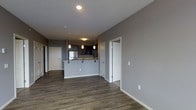 This single-floor space showcases a thoughtfully designed open-concept layout that seamlessly connects the living area with the kitchen. The kitchen is a standout feature, anchored by a raised peninsula with a light-toned countertop that provides both additional prep space and casual seating. Dark cabinetry adds a bold contrast to the lighter surroundings, while stainless steel appliances and pendant lighting lend a polished, contemporary feel. A track lighting system overhead further enhances the kitchen's functionality and style. Rich, dark hardwood-style flooring runs throughout the space, grounding the neutral gray walls and white trim in a warm, cohesive palette. The open living area benefits from the flowing layout, offering a versatile and spacious feel. Multiple doorways lead to additional rooms, suggesting a well-organized floor plan with distinct private spaces accessible from the main living area. Recessed lighting in the ceiling provides clean, even illumination. Overall, the space presents a modern, practical, and aesthetically cohesive environment with quality finishes that balance style and everyday functionality.