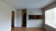 This single-floor space offers a straightforward and functional layout designed to maximize usability. The living area flows seamlessly toward the kitchen, which is accessible via a pass-through counter finished with a dark granite-style surface — a practical and visually appealing feature that connects the two spaces while maintaining a sense of separation. The kitchen is equipped with black appliances, including a refrigerator and range, set against dark cabinetry and an under-cabinet range hood, giving the cooking area a cohesive, modern look. Rich, dark hardwood-style flooring runs throughout the main living areas, adding warmth and a clean, polished aesthetic. The walls are finished in a neutral light gray, providing a versatile backdrop that complements a variety of furnishing styles. A textured popcorn ceiling is consistent throughout the space. A hallway visible from the main area leads to additional rooms, with doors providing privacy to each. Overhead HVAC vents are integrated into the ceiling, ensuring climate control throughout. The overall ambiance is compact yet well-organized, with a layout that makes efficient use of every area.