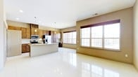 This single-floor space showcases a thoughtfully designed open-concept layout that seamlessly connects the kitchen and living areas. The kitchen is well-appointed with dark wood cabinetry, stainless steel appliances including a microwave and French door refrigerator, and granite countertops. A central kitchen island provides additional prep space and serves as a natural dividing point between the culinary and living zones. A built-in wine rack adds a distinctive architectural touch above the upper cabinets. Pendant lighting fixtures hang above the island, complementing the recessed lighting throughout the ceiling to create a warm, layered lighting scheme. The living area is expansive and open, finished with large-format, high-gloss white tile flooring that reflects light beautifully across the space. Oversized windows with grid detailing line the walls, inviting generous amounts of natural light and offering views of the surrounding neighborhood. The warm taupe wall tones contrast elegantly with the bright flooring and light ceiling, lending the space a balanced, neutral palette. Overall, the space offers a clean, modern aesthetic with functional design elements suited for both everyday living and entertaining.