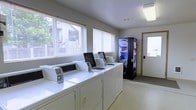 This single-floor communal laundry facility is designed with functionality and convenience in mind. The room is bright and welcoming, with multiple large windows fitted with horizontal blinds that allow generous natural light to fill the space while maintaining privacy. A row of top-loading washing machines lines one side of the room, paired with matching dryers, providing ample capacity to serve multiple users simultaneously. The machines are arranged in an orderly fashion, making the space easy to navigate. A Pepsi vending machine is conveniently positioned near the entrance, offering refreshments while residents wait. The room features a clean, neutral color palette with light-colored walls and flooring that contributes to a tidy, utilitarian atmosphere. A standard entry door with a secure keypad lock provides controlled access to the facility, enhancing the safety and security of the space. Overhead fluorescent lighting supplements the natural light, ensuring the room remains well-lit at all hours. The overall layout is practical and efficiently organized, making this laundry room a fully functional and accessible shared amenity.