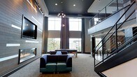 This sophisticated two-story communal lounge is designed to impress with its dramatic double-height ceilings and an expansive, open atmosphere. The space is anchored by a striking dark-painted ceiling that contrasts beautifully with the light-toned flooring and neutral walls, creating a bold yet inviting aesthetic.

The main level offers multiple seating arrangements, including deep, cushioned sofas and armchairs in rich jewel tones — perfect for relaxed conversation or lounging. A contemporary fireplace is built into the feature wall alongside a large flat-screen television, adding warmth and a focal point to the room. A billiards table occupies a central position, providing a natural gathering spot for recreation.

Floor-to-ceiling windows flood the interior with abundant natural light and offer open views of the surrounding landscape, enhancing the airy feel of the space. Elegant pendant lighting and recessed fixtures provide ambient illumination throughout.

A sleek staircase with modern cable railings connects the main level to an open upper mezzanine, which overlooks the lounge below. The mezzanine level appears to offer additional seating and activity areas, extending the usable space vertically. The overall design reflects a contemporary, upscale sensibility with clean lines, thoughtful material choices, and a cohesive dark-and-neutral color palette.