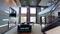This sophisticated two-story communal lounge is designed to impress with its dramatic double-height ceilings and an expansive, open atmosphere. The space is anchored by a striking dark-painted ceiling that contrasts beautifully with the light-toned flooring and neutral walls, creating a bold yet inviting aesthetic.

The main level offers multiple seating arrangements, including deep, cushioned sofas and armchairs in rich jewel tones — perfect for relaxed conversation or lounging. A contemporary fireplace is built into the feature wall alongside a large flat-screen television, adding warmth and a focal point to the room. A billiards table occupies a central position, providing a natural gathering spot for recreation.

Floor-to-ceiling windows flood the interior with abundant natural light and offer open views of the surrounding landscape, enhancing the airy feel of the space. Elegant pendant lighting and recessed fixtures provide ambient illumination throughout.

A sleek staircase with modern cable railings connects the main level to an open upper mezzanine, which overlooks the lounge below. The mezzanine level appears to offer additional seating and activity areas, extending the usable space vertically. The overall design reflects a contemporary, upscale sensibility with clean lines, thoughtful material choices, and a cohesive dark-and-neutral color palette.