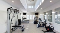 This fitness center is designed to offer a bright, open, and motivating environment for exercise. The space features soaring two-story ceilings with clerestory windows positioned high on the walls, flooding the room with natural light and creating an airy, spacious atmosphere. A ceiling fan overhead aids in air circulation, contributing to a comfortable workout environment.

Large windows along the lower walls provide additional natural light and views to the outdoors, while recessed lighting throughout ensures the space remains well-lit at all hours. Wall-mounted flat-screen televisions are positioned for easy viewing from multiple workout stations.

The fitness center is comprehensively equipped for both cardio and strength training. Cardio equipment includes treadmills, an elliptical trainer, and a recumbent bike, all from Matrix, a reputable fitness brand. A dedicated free weights area features a full rack of dumbbells alongside an adjustable weight bench, catering to a variety of strength training routines. A multi-function cable and resistance machine further expands the strength training options available.

Full-length mirrors are positioned along one wall, allowing users to monitor their form during workouts. The flooring is a durable, low-pile carpet suitable for a fitness setting. The clean, neutral color palette of white walls and light tones throughout contributes to a fresh and professional ambiance.