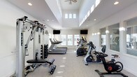 This fitness center is designed to offer a bright, open, and motivating environment for exercise. The space features soaring two-story ceilings with clerestory windows positioned high on the walls, flooding the room with natural light and creating an airy, spacious atmosphere. A ceiling fan overhead aids in air circulation, contributing to a comfortable workout environment.

Large windows along the lower walls provide additional natural light and views to the outdoors, while recessed lighting throughout ensures the space remains well-lit at all hours. Wall-mounted flat-screen televisions are positioned for easy viewing from multiple workout stations.

The fitness center is comprehensively equipped for both cardio and strength training. Cardio equipment includes treadmills, an elliptical trainer, and a recumbent bike, all from Matrix, a reputable fitness brand. A dedicated free weights area features a full rack of dumbbells alongside an adjustable weight bench, catering to a variety of strength training routines. A multi-function cable and resistance machine further expands the strength training options available.

Full-length mirrors are positioned along one wall, allowing users to monitor their form during workouts. The flooring is a durable, low-pile carpet suitable for a fitness setting. The clean, neutral color palette of white walls and light tones throughout contributes to a fresh and professional ambiance.